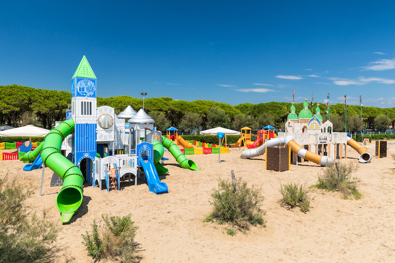 Colorful playground structures with slides in a sandy area surrounded by greenery and trees at Residence Village