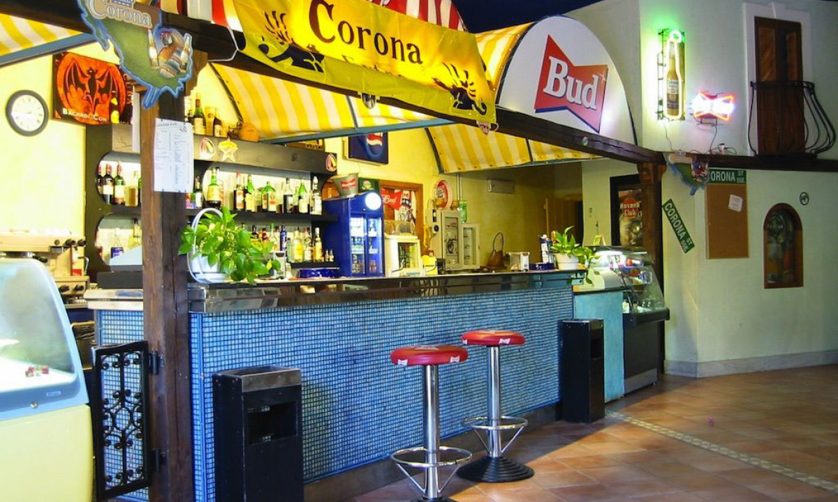 At Ultima Spiaggia, a bar counter featuring two crimson stools is stocked with bottles and adorned with decorations, all situated within a well-lit camping area.
