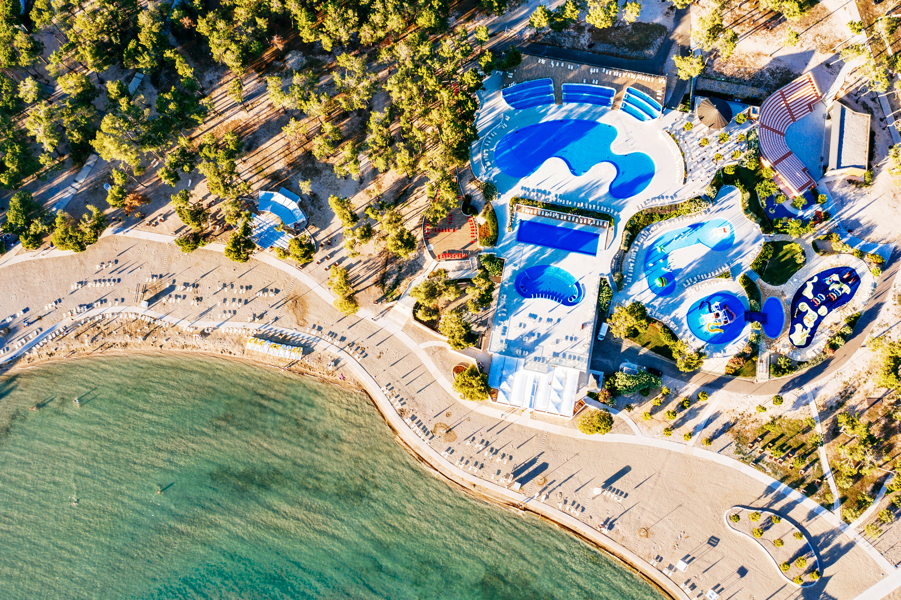 At Zaton Holiday Resort, there are swimming pools featuring blue slides, encircled by trees and a sandy beach adjacent to a tranquil shoreline.