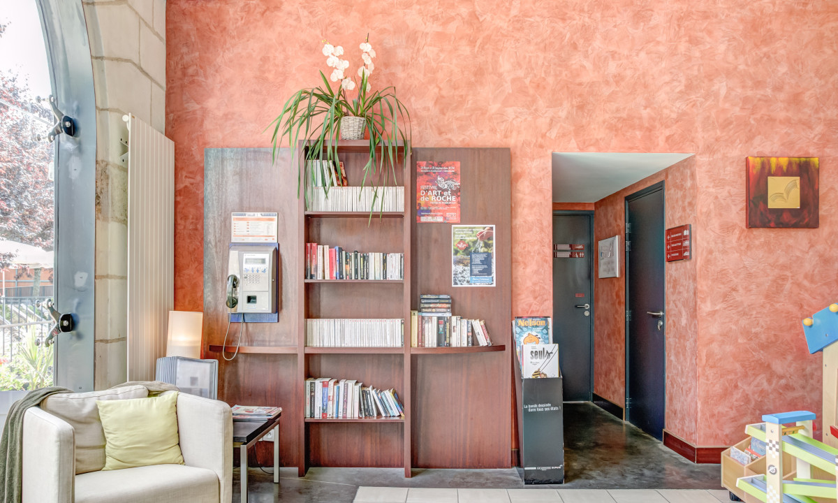 Bookshelf holding books, magazines, and pamphlets next to a cozy armchair by a large window; indoors library scene at Le Moulin des Cordeliers