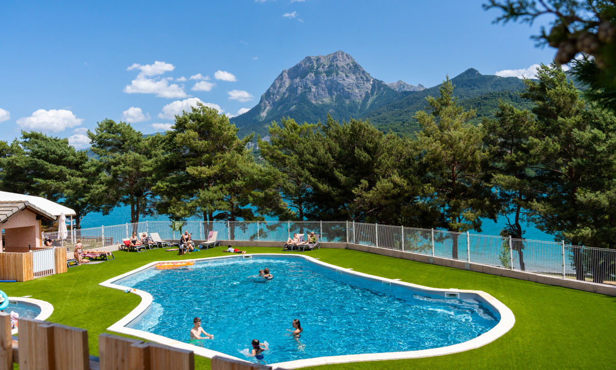 At La Presqu’île Serre-Ponçon, a swimming pool with individuals unwinding is encircled by trees and mountains near a lake.