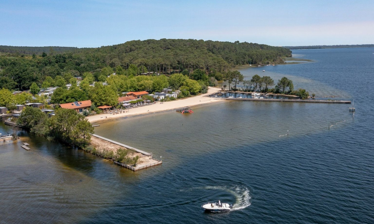A boat moves on a lake near a sandy beach, surrounded by trees and cabins at Maguide