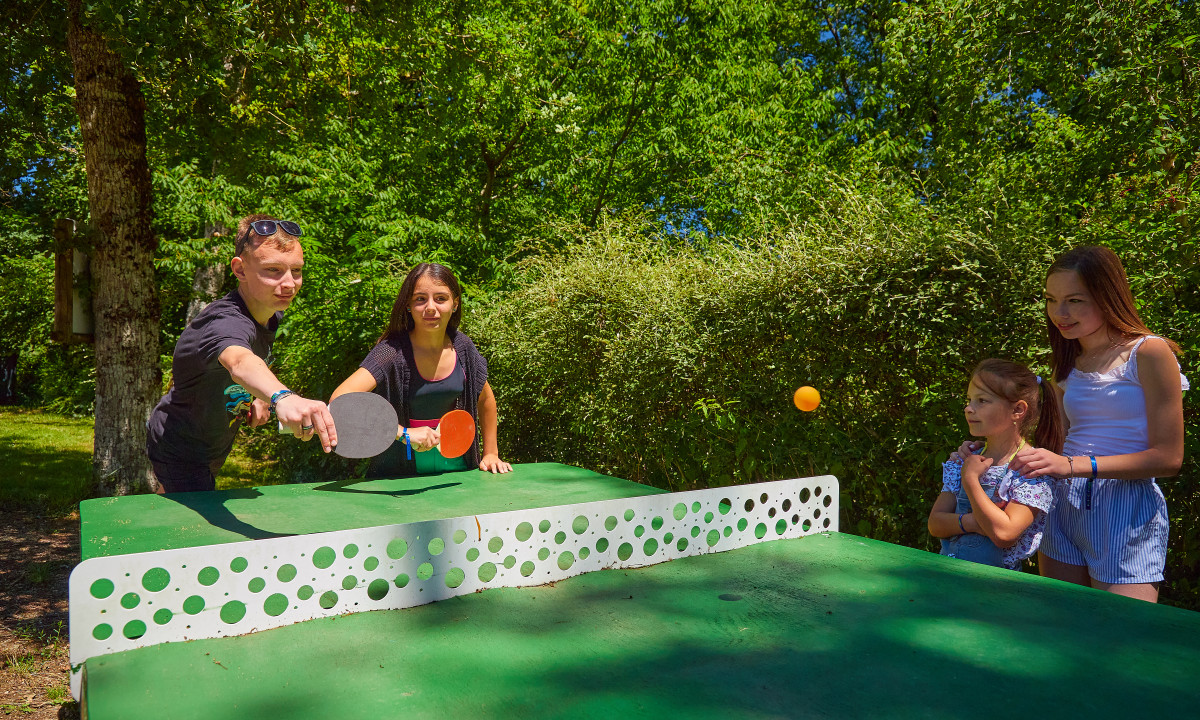 At Terrasses du Lac, two individuals engage in table tennis while two others observe, encircled by vibrant foliage.