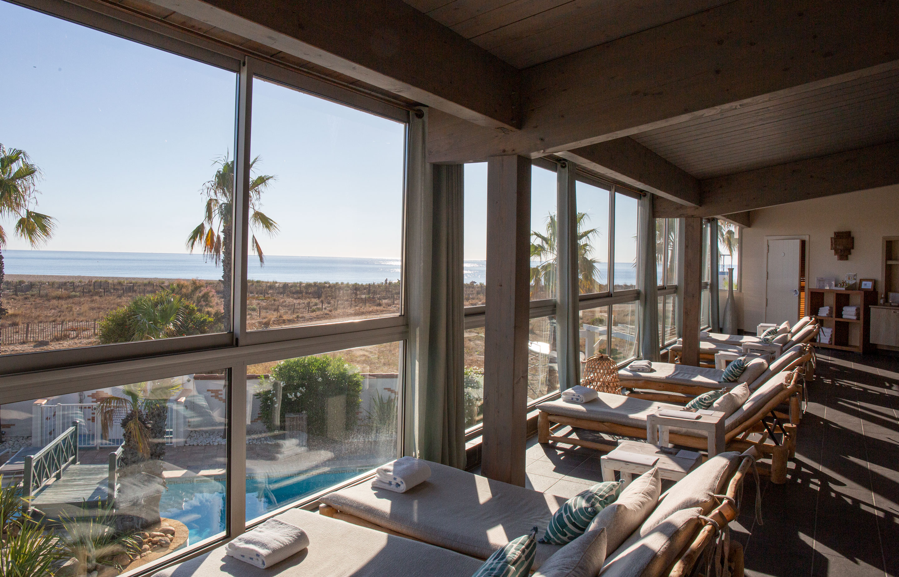 Lounge chairs beside large windows face the ocean, overlooking a pool with palm trees visible outside at Les Bulles de Mer