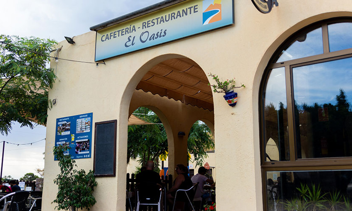 At Camping Los Escullos, the entrance of a café features patrons seated beneath arches, enveloped by trees and a verdant outdoor environment.