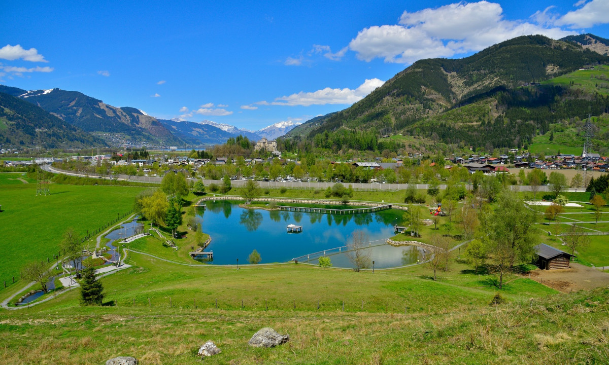 A tranquil pond reflecting mountains and trees, surrounded by green fields and a small village, at Sportcamp Woferlgut