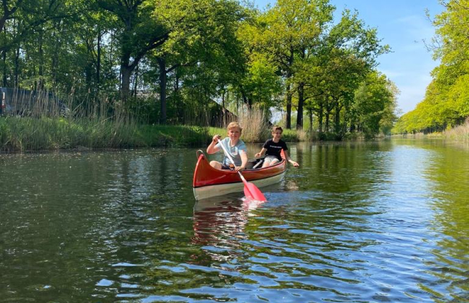 At de Vinkenkamp, two individuals paddle a crimson canoe along a tree-fringed river, relishing a sunny day.