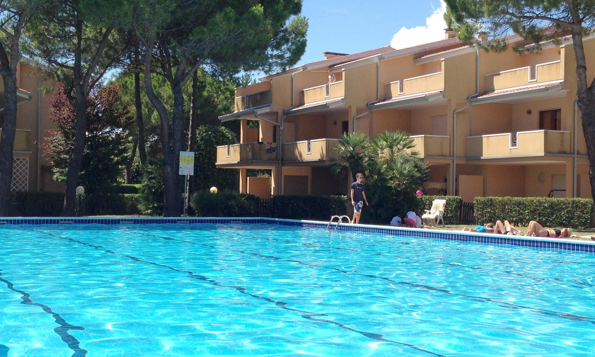 Swimming pool reflecting sunlight with people sunbathing, located near a yellow apartment complex surrounded by trees at Villaggio Selene