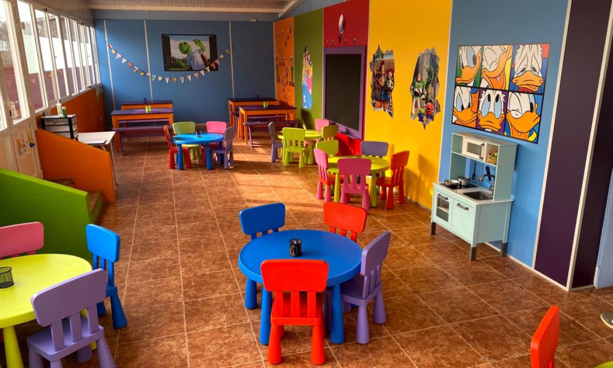 Colorful children's tables and chairs arranged in a bright playroom with cartoon decorations on walls and tiled floor at Resort Els Pins