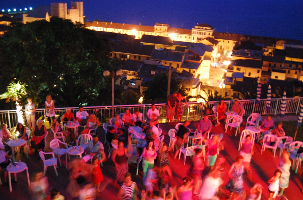 People sitting and dancing on a terrace with tables and chairs, overlooking a lit town at night at Sant' Anna Residence