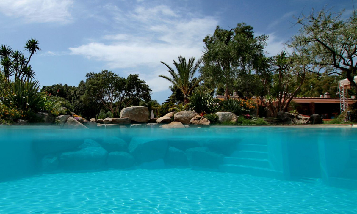 At Ultima Spiaggia, a pool filled with transparent water is surrounded by stones and vibrant greenery, with a bright sky overhead.
