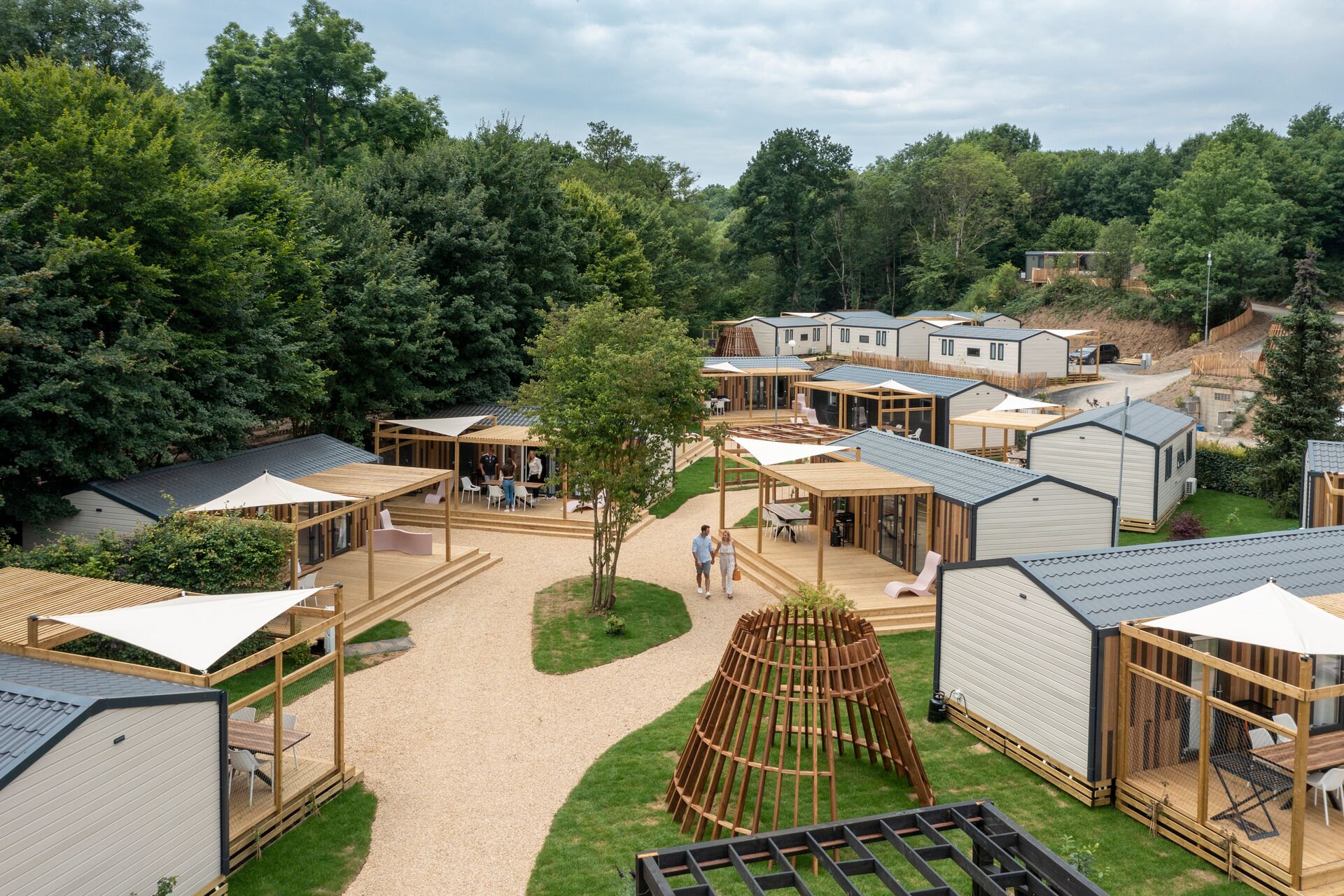 At Parc La Clusure, individuals stroll among contemporary mobile residences featuring wooden patios, encircled by abundant trees and greenery.
