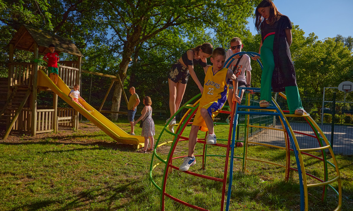 At Terrasses du Lac, youngsters ascend play structures and glide down alongside adults in a sunlit park environment.