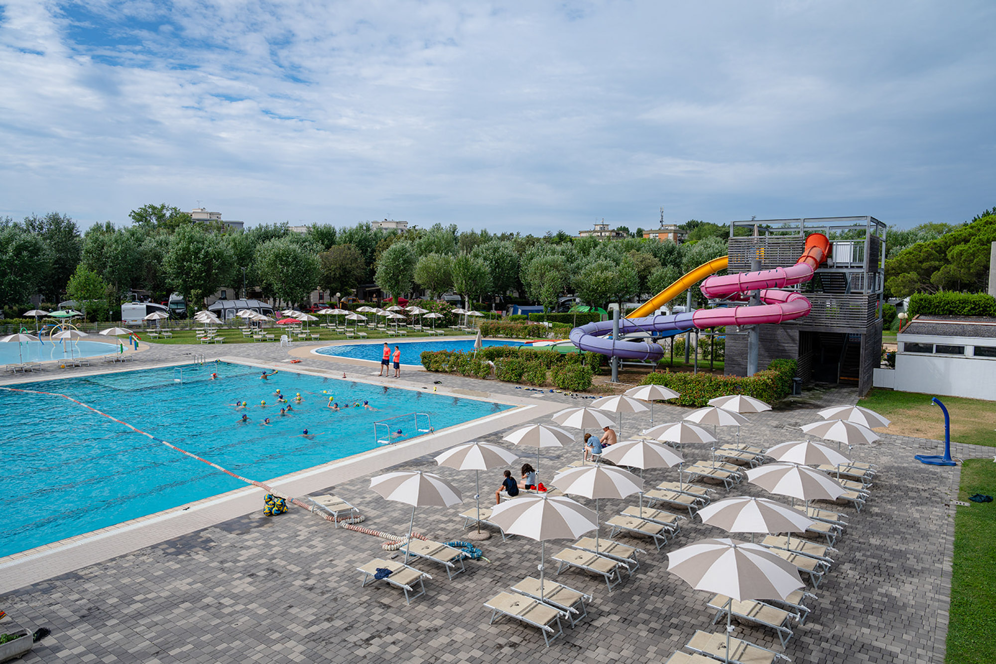 Swimming pool with people playing; nearby water slides and sun loungers; surrounded by trees and umbrellas at Marina Julia Family Camping Village