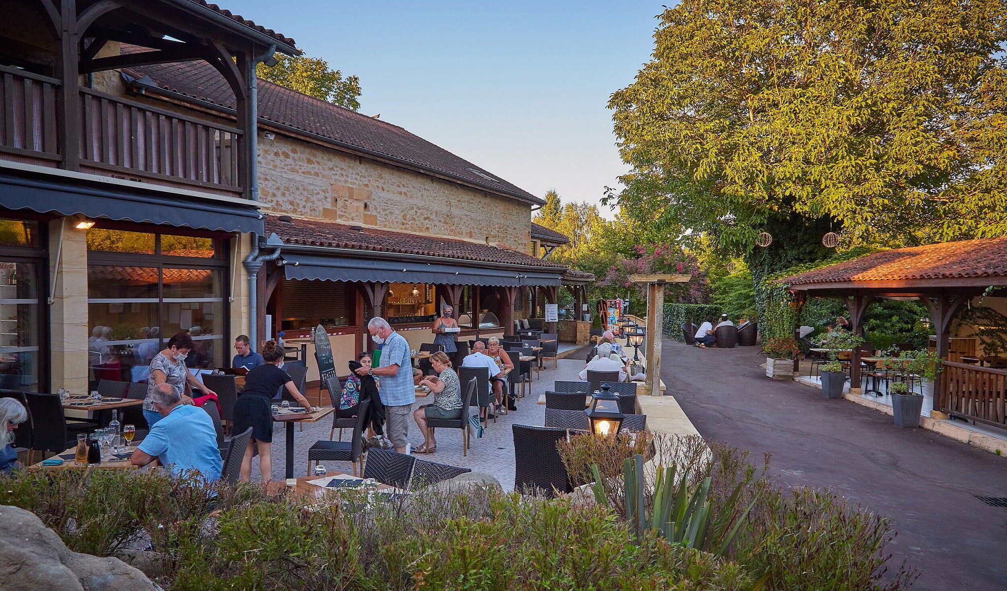 At Marvilla Parks Domaine de Soleil Plage, individuals are enjoying meals at alfresco tables next to a charming stone structure, enveloped by lush vegetation in a laid-back ambiance.