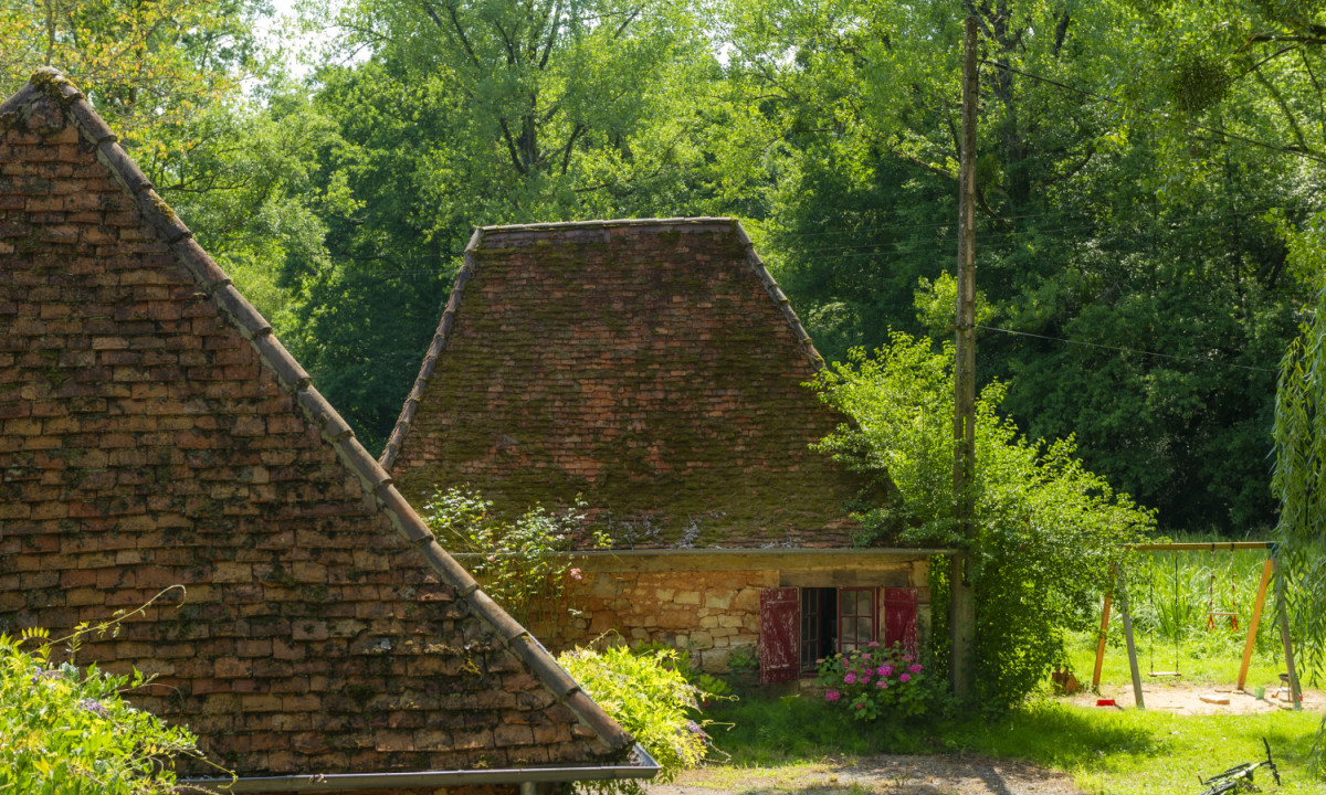 At Coucou, an ancient stone dwelling features a brick roof surrounded by vibrant foliage and trees, with playground apparatus visible in the background.