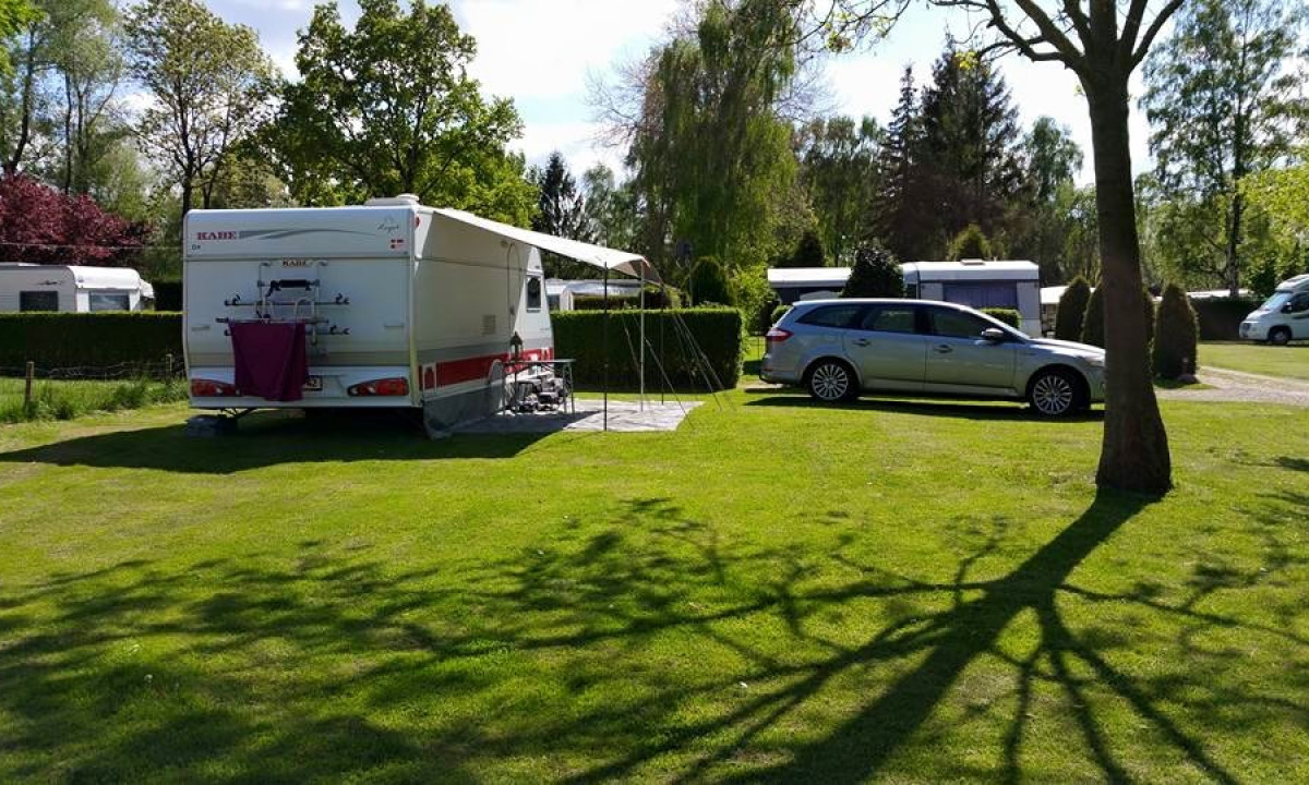 At Elbeling, a stationary caravan with an extended canopy is positioned in a sunny, grassy campsite close to trees and other vehicles.