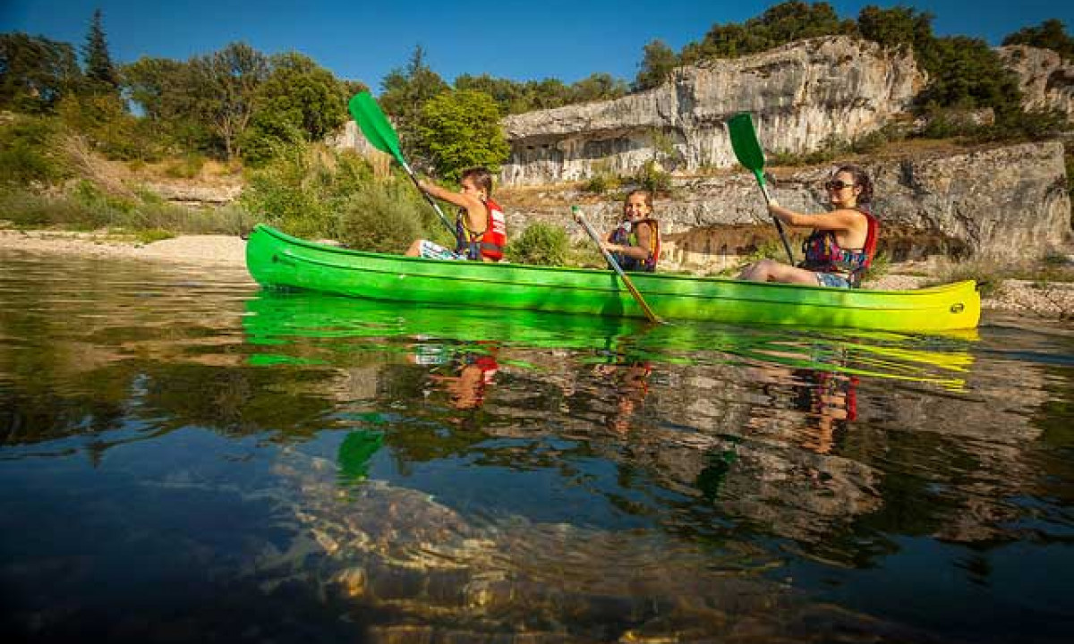 At La Sousta, three individuals paddle a green kayak on a tranquil river, with rugged cliffs and lush vegetation in the backdrop.