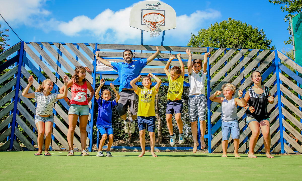 Nine people jumping on a grassy sports court, posing in front of a blue wooden basketball hoop structure at Atlantica