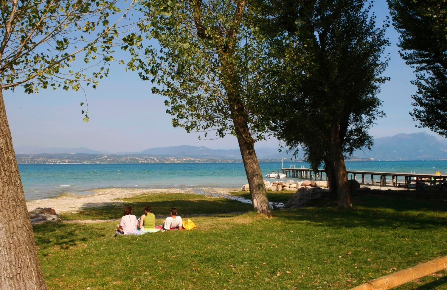 Three people sit on grass under trees, facing a lake with a pier, mountains in the background at San Francesco