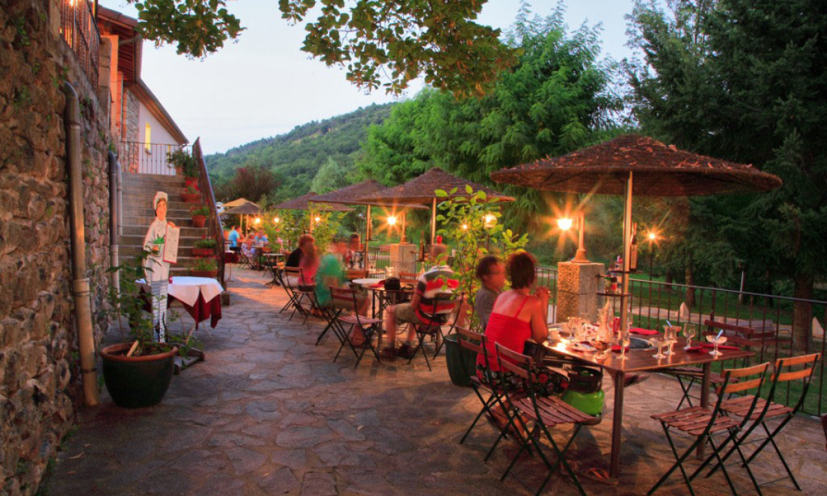 At Les Ranchisses, individuals are enjoying meals at tables outside under umbrellas, encircled by trees and a charming stone structure.