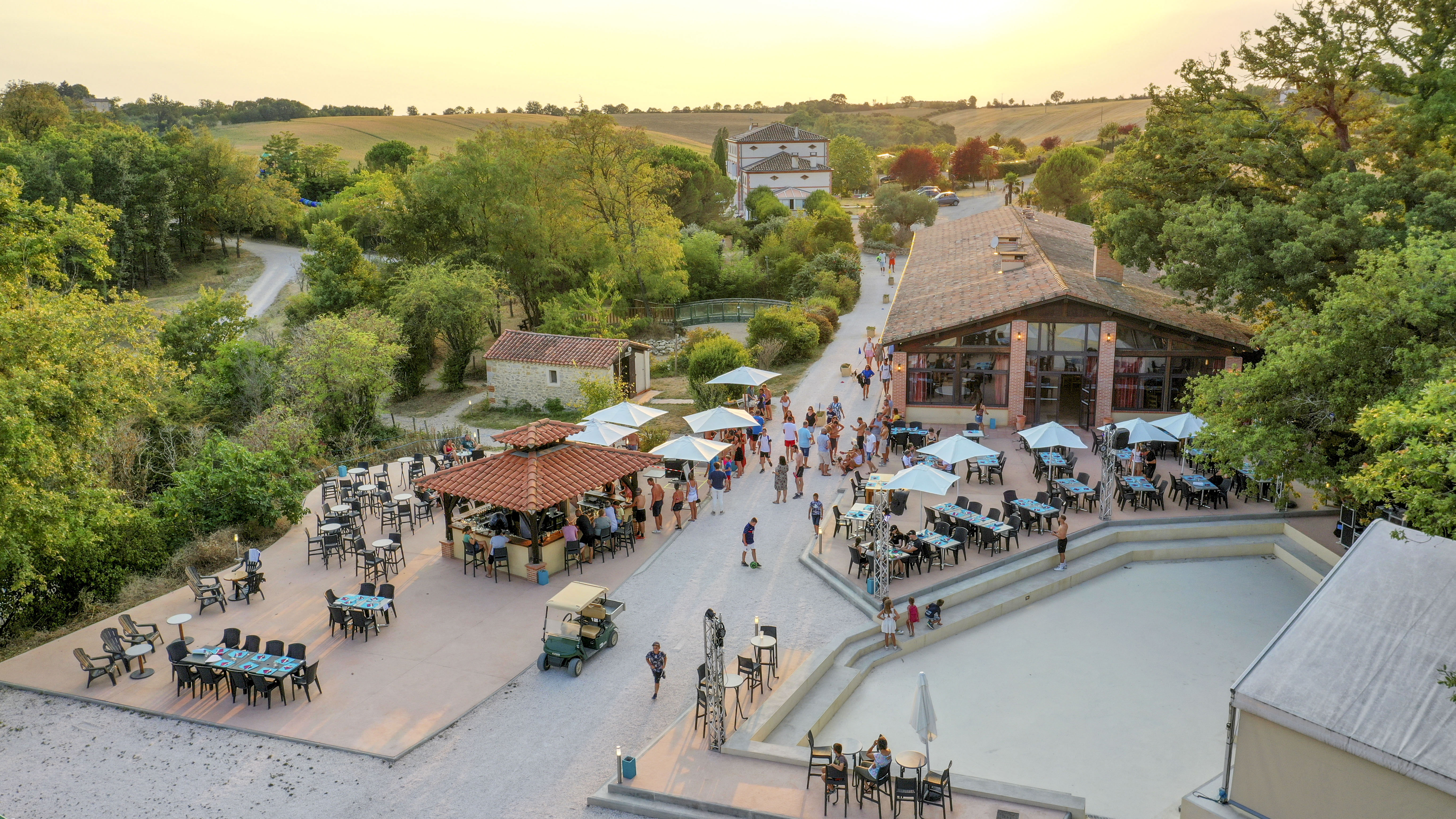 At Domaine du Koukano, individuals are enjoying meals and interacting in an outdoor seating space surrounded by large trees and a nearby structure.
