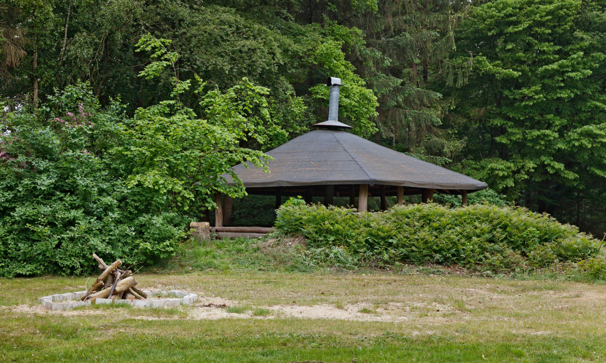At KNAUS Campingpark Wingst, a timber shelter featuring a conical roof is situated within a thick forest, next to a campfire area with unlit logs.