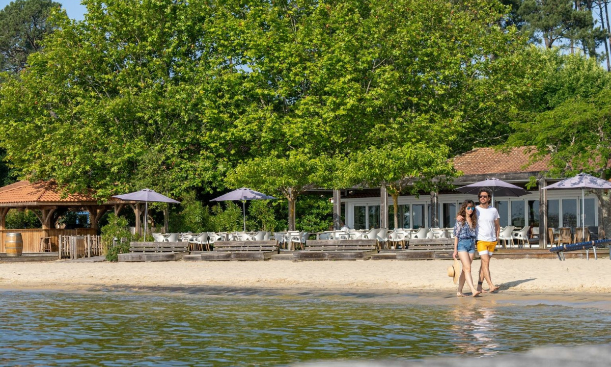 A couple walks along the sandy shoreline near a beachside café with leafy trees at Maguide