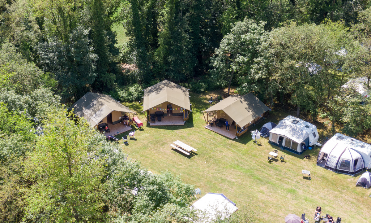 Several tents and caravans surrounded by trees with people sitting and socializing on a grassy area at de Wrange