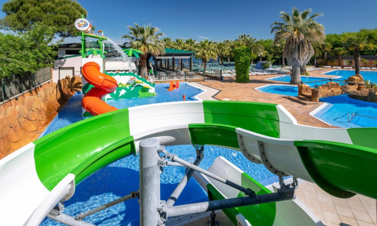 A green waterslide curves above a pool area with palm trees and lounge chairs, creating a vibrant aquatic setting at Solmar