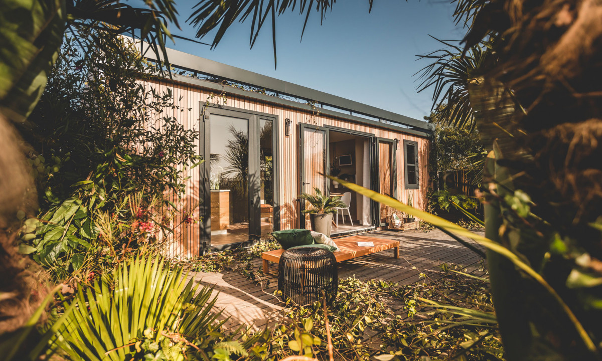 At La Clémentine, a contemporary cabin with open entrances is enveloped by vibrant foliage and sunlight, forming a warm outdoor area.