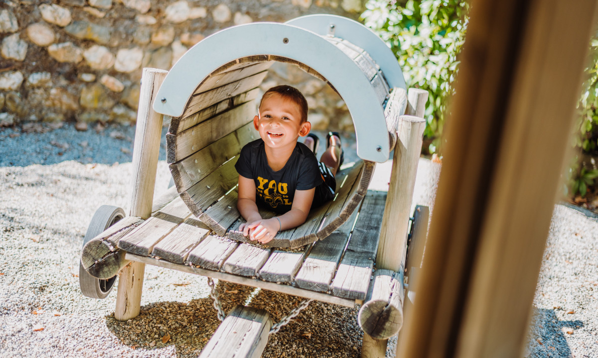 At La Clémentine, a child is reclining inside a wooden play tunnel, grinning, with a stone wall and lush greenery in the background.