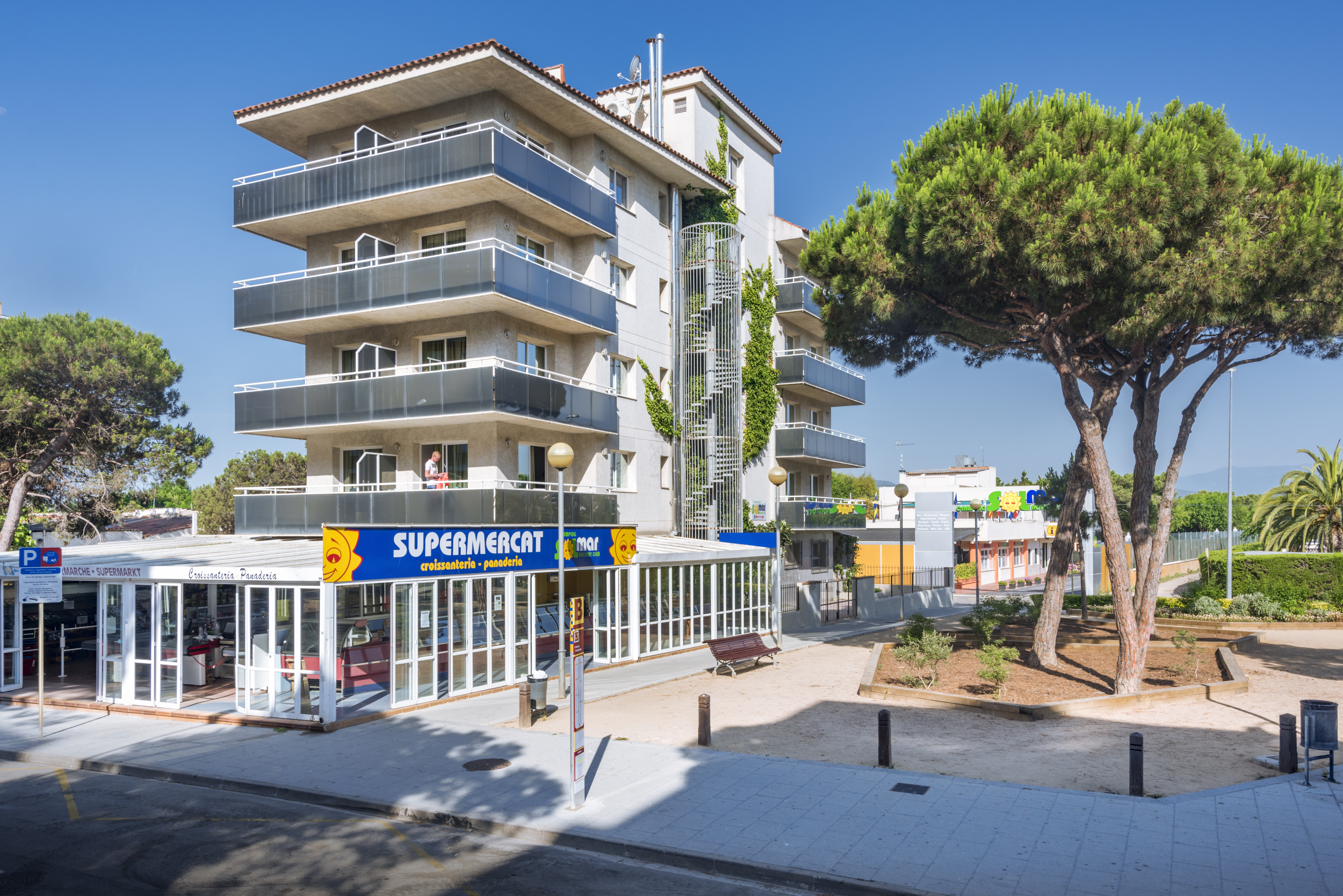 At Solmar, there is a four-level structure featuring terraces, tall pine trees, and a grocery store on the first floor, along with an urban walkway and road.