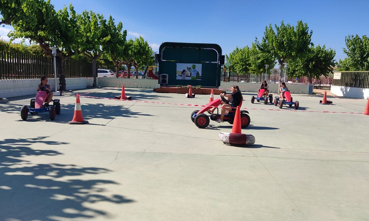 Children riding pedal go-karts on a paved track with traffic cones, under the shade of green trees at Resort Els Pins