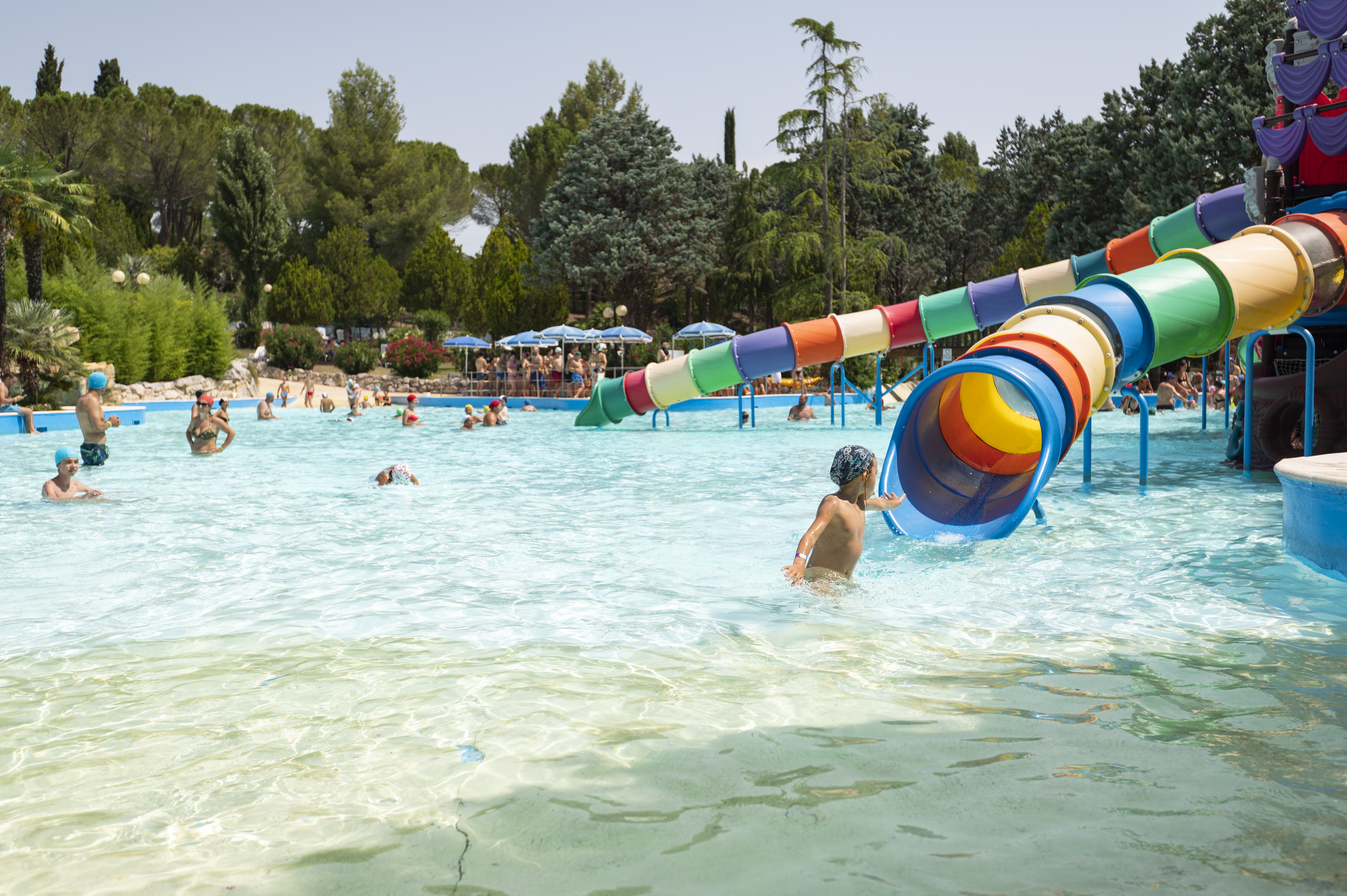 Children playing in a water park pool with a colorful slide, surrounded by trees and people lounging at Centro Vacanze Verde Azzurro