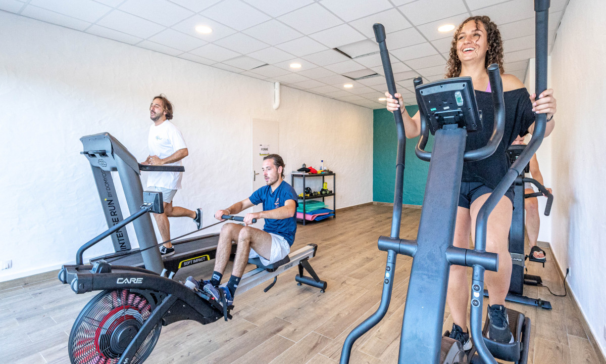 People exercising on gym equipment in a brightly lit indoor gym with white walls and wooden floors at Atlantica