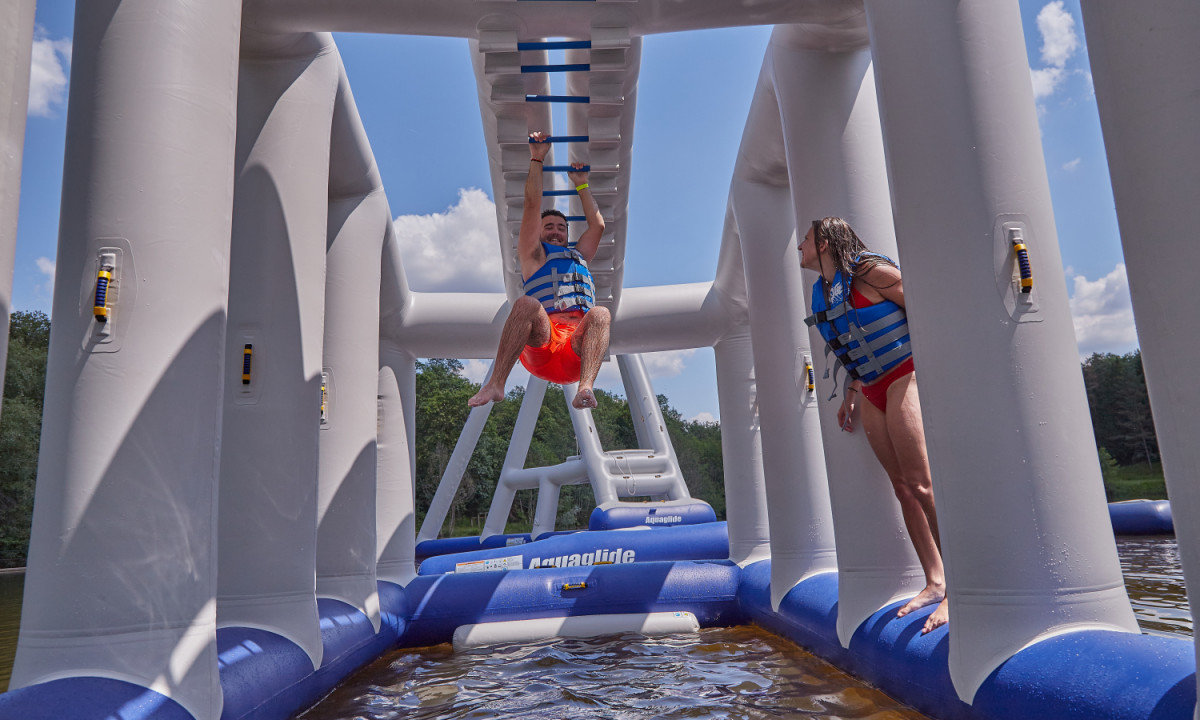 At Coucou, an individual ascends a buoyant challenge course while another remains on standby, both donning life jackets above the water.