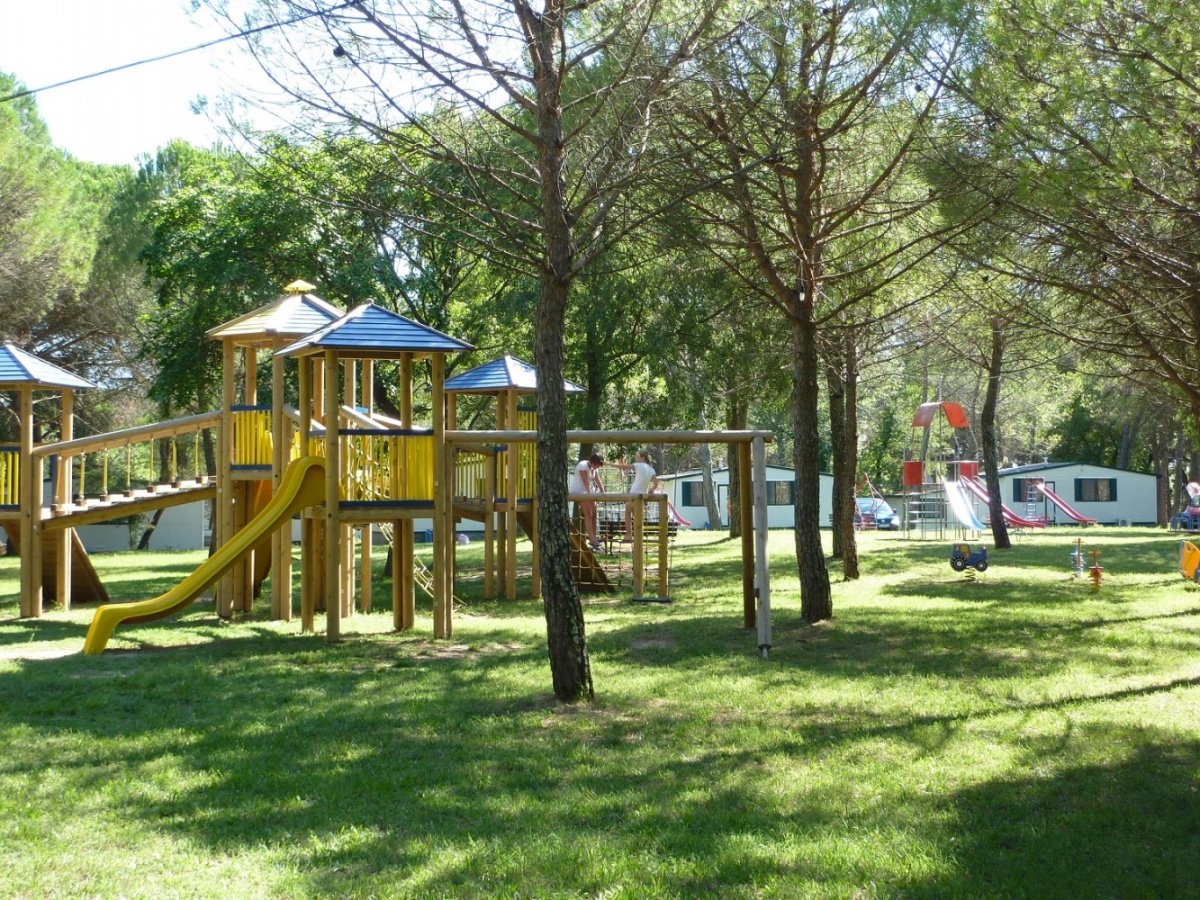 Children's playground equipment with slides and bridges among tall trees, mobile homes visible in the background at Belvedere Pineta