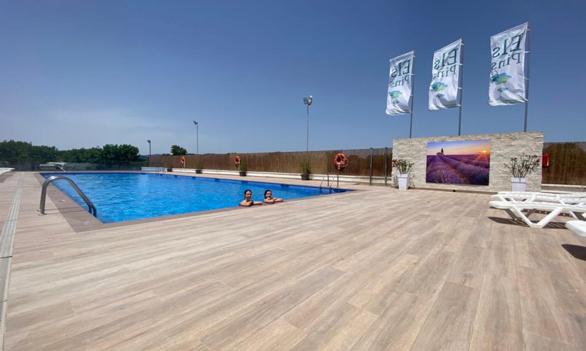 Swimming pool with two people in the water, surrounded by a spacious deck, flags, and trees in the background at Resort Els Pins