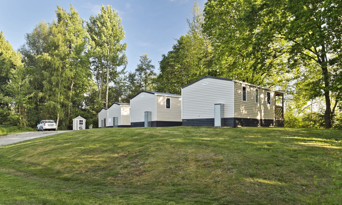 At KNAUS Campingpark Oyten, three small white cottages are positioned next to each other on a grassy area, with a parked vehicle nearby, encircled by tall green trees.