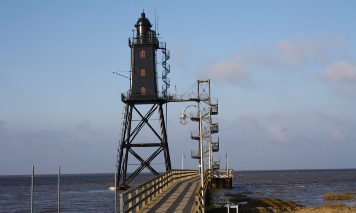 At KNAUS Campingpark Dorum, a dark-hued lighthouse rises prominently on a jetty, gazing over the tranquil ocean beneath a clear sky.