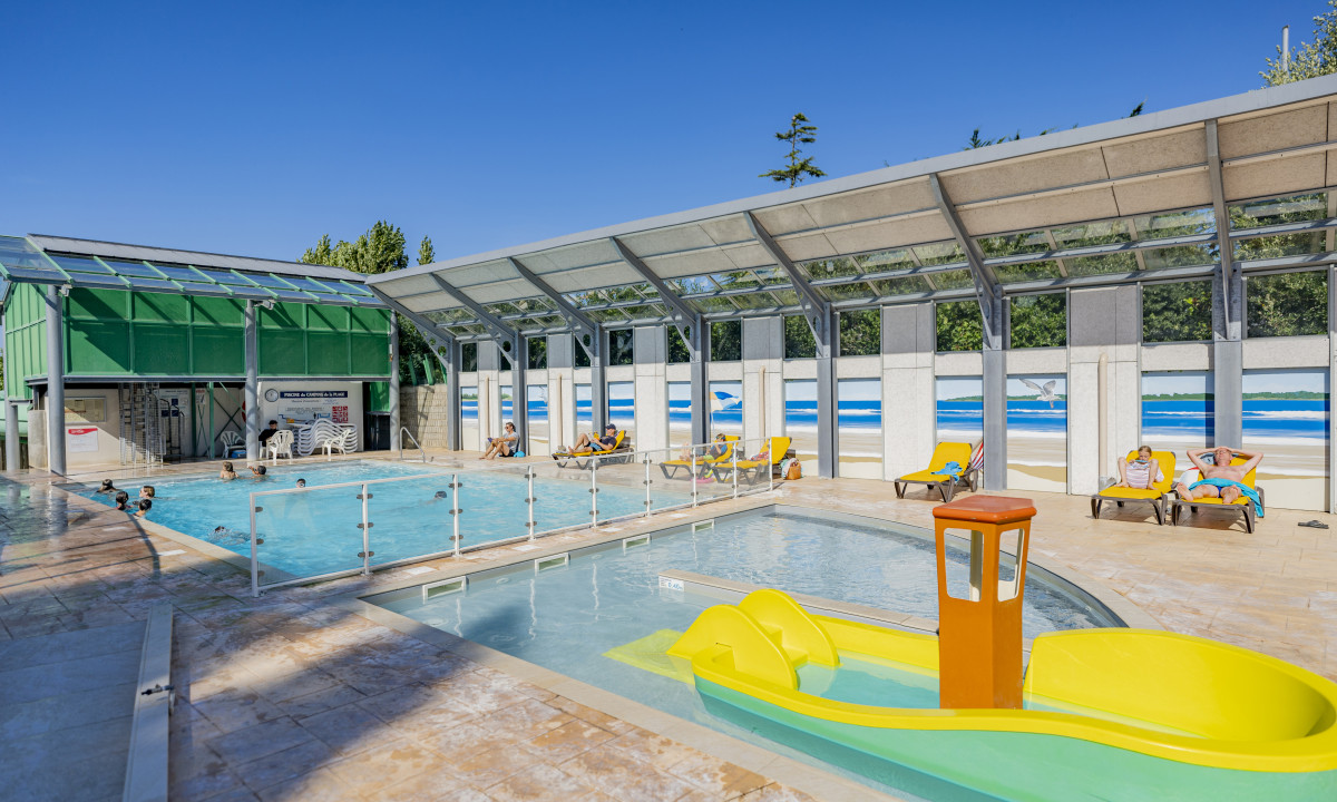 At De la Plage, kids are swimming in a pool with a slide, encircled by sunbathers on lounge chairs beneath a clear sky.