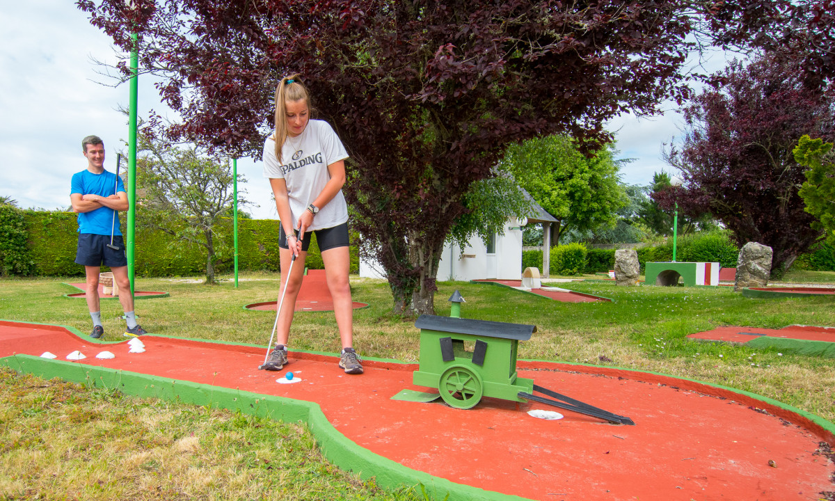 At De la Plage, an individual is playing mini-golf while another observes, encircled by trees and ornamental obstacles in a park environment.