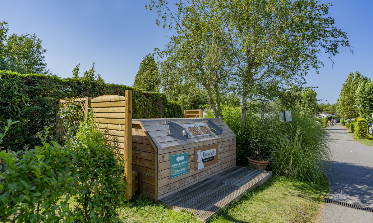 At De la Plage, a wooden recycling container is situated amidst vibrant foliage along a sunlit path.