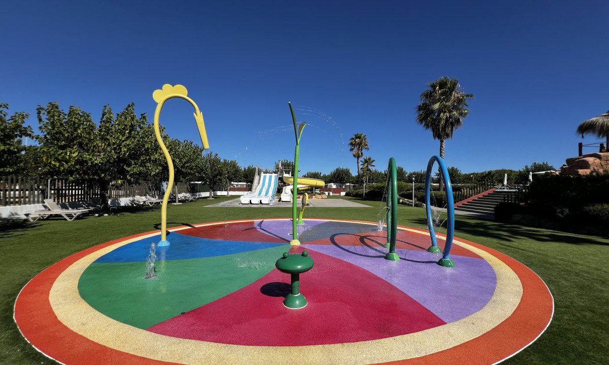 Colorful splash pad with water jets spraying, surrounded by trees and playground slides at Resort Els Pins
