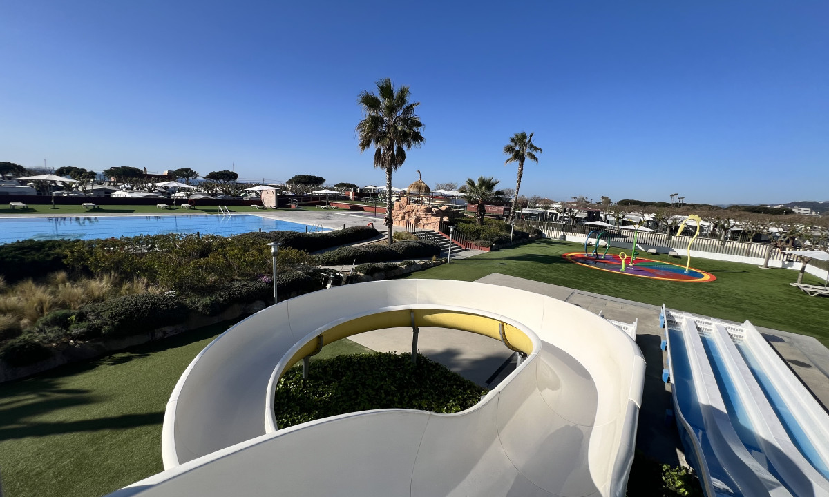 Curved waterslide overlooks a large pool and palm trees, surrounded by greenery and a clear blue sky at Resort Els Pins