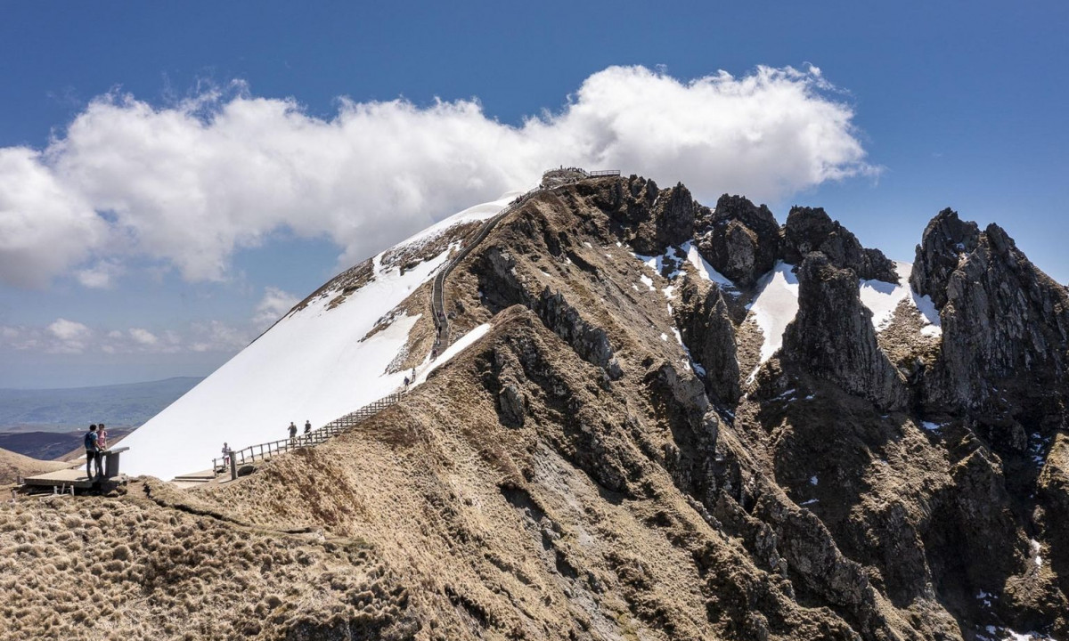 Hikers walking along a fenced mountain ridge with snow patches; cloudy, blue sky backdrop at La Ribeyre