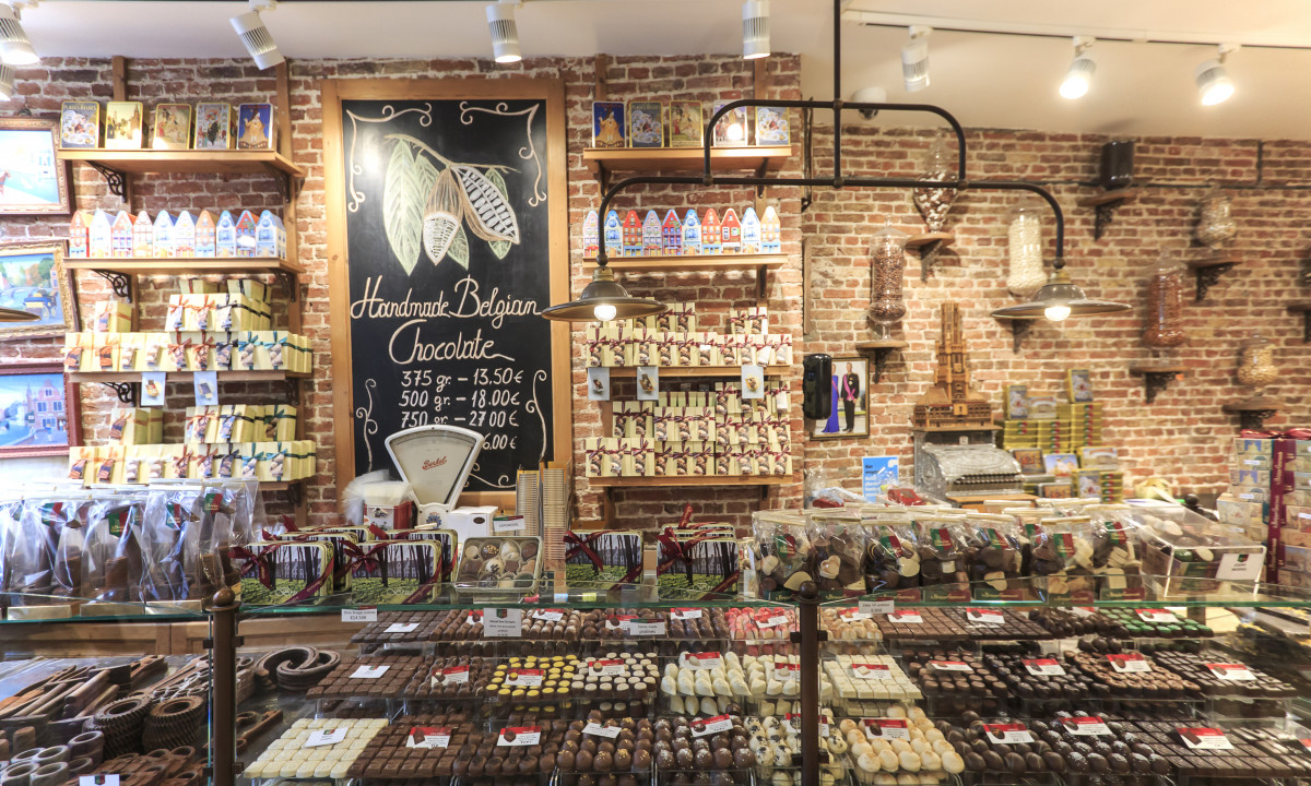 At Urban Gardens Gent, shelves showcase an assortment of packaged chocolates behind a counter, while boxed chocolates are presented under glass in a brick-walled store.