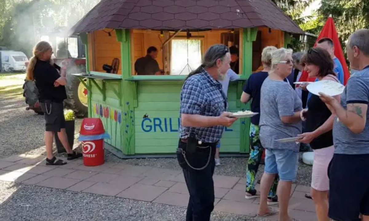 At Elbeling, individuals with dishes are queued at a green grill stand located within a forested camping area.