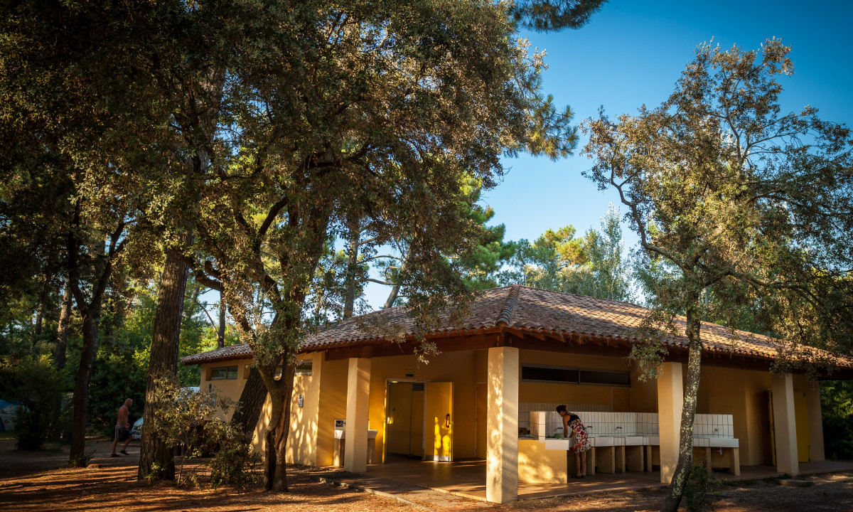 At La Sousta, an individual cleans dishes outside a yellow structure encircled by trees in a wooded camping area.